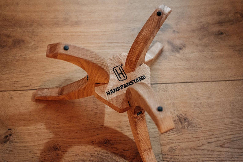 May include: A wooden handpan stand with a hexagonal base and four legs. The stand is engraved with the text "HANDPANSTAND" and a stylised "H".