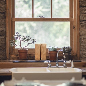Peut inclure: Un &eacute;vier de cuisine en porcelaine blanche avec un robinet chrom&eacute;. L'&eacute;vier est pos&eacute; sur un plan de travail en bois rustique et un mur en brique. Il y a des plantes en pot et des blocs de bois sur le rebord de la fen&ecirc;tre derri&egrave;re l'&eacute;vier.