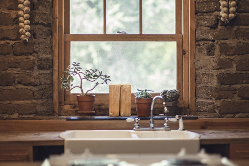 Peut inclure: Un &eacute;vier de cuisine en porcelaine blanche avec un robinet chrom&eacute;. L'&eacute;vier est pos&eacute; sur un plan de travail en bois rustique et un mur en brique. Il y a des plantes en pot et des blocs de bois sur le rebord de la fen&ecirc;tre derri&egrave;re l'&eacute;vier.