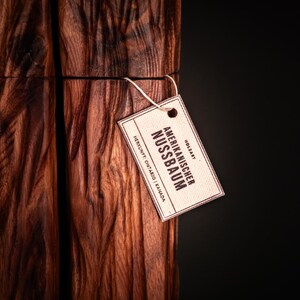 Op de afbeelding: Close-up van een houten object met een diepe bruine kleur en ingewikkelde nerfpatronen. Een klein label is bevestigd, met de tekst "Amerikanischer Nussbaum". De achtergrond is effen zwart.