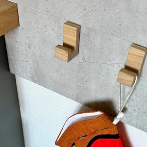 May include: Two wooden coat hooks mounted on a gray wall. The hooks are a light brown color and have a simple, modern design. One hook holds a decorative item with red and orange fabric.