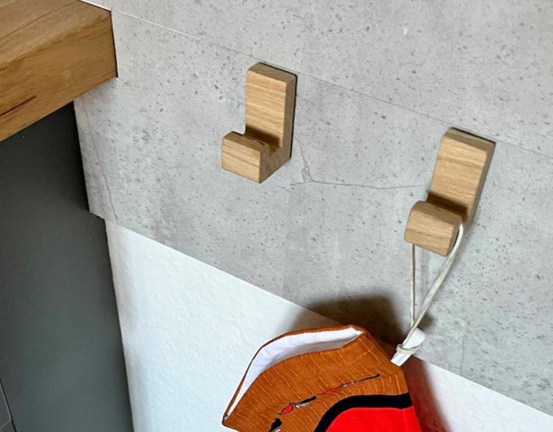 May include: Two wooden coat hooks mounted on a gray wall. The hooks are a light brown color and have a simple, modern design. One hook holds a decorative item with red and orange fabric.