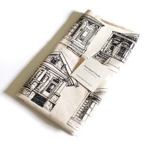 May include: A cream-colored tea towel with a black and white screen-printed design of Victorian-style houses. The towel has a tag that reads "Statement Goods by The Collective Shop LLC. Screen-Printed Tea Towel."