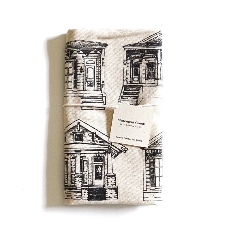 May include: A cream-colored tea towel with a black and white illustration of four houses. The towel has a tag that reads "Statement Goods by The Collective Shop LLC. Screen-Printed Tea Towel."