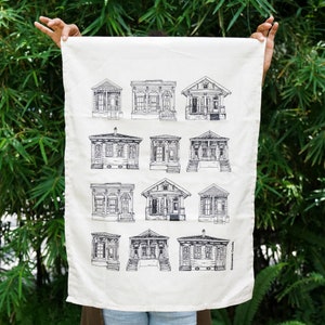 May include: A white kitchen towel with a black and white illustration of 15 different New Orleans style houses. The text "Statement Goods" is printed in the bottom right corner of the towel.