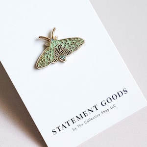 May include: A gold-toned moth pin with a mint green and gold design, resting on a white card. The pin features intricate details and is set against a clean background. The card reads "STATEMENT GOODS" in black text.