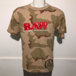 Op de afbeelding: Een camouflageprint t-shirt met het woord "RAW" in rode letters en de tekst "Natural Unrefined Rolling Papers" in witte letters. Het shirt heeft ook een klein bruin patch met de tekst "Authentic Purest Natural Hemp Fibers Unrefined".