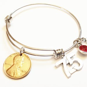 May include: Silver-toned bangle bracelet with a gold-colored penny charm, a silver number 75 charm, a red gemstone charm, and a silver number 5 charm. The bracelet is on a white background.