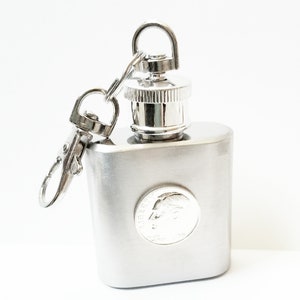 May include: A silver flask with a coin design on the front and a screw-on cap. The flask has a silver chain attached to it.