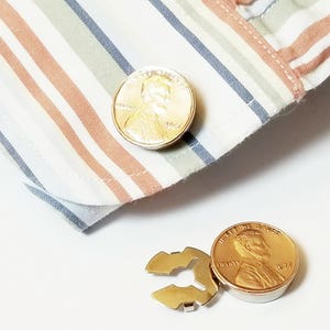May include: A pair of gold-colored cufflinks featuring a design of a penny with the profile of Abraham Lincoln. The cufflinks are on a striped fabric background.