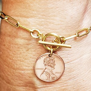 Puede incluir: Pulsera de cadena dorada con cierre de palanca dorado y un dije de centavo estadounidense de 2022. El centavo presenta un perfil de Abraham Lincoln y las palabras "In God We Trust" y "Liberty".