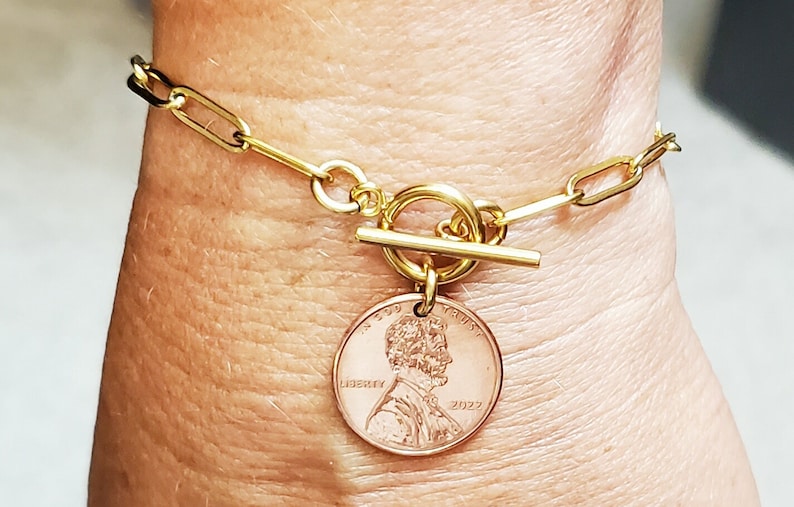 Puede incluir: Pulsera de cadena dorada con cierre de palanca dorado y un dije de centavo estadounidense de 2022. El centavo presenta un perfil de Abraham Lincoln y las palabras "In God We Trust" y "Liberty".