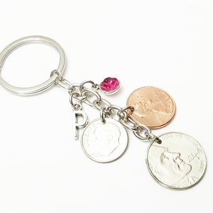 May include: A silver keyring with a pink rhinestone charm, a silver letter 'P' charm, and three US coins: a penny, a nickel, and a dime. The penny is dated 2005 and the dime is dated 1964.