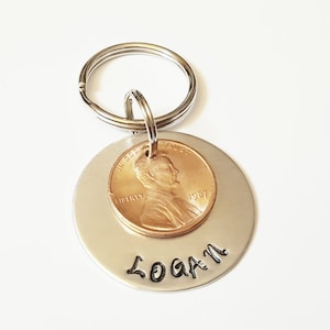 May include: Silver key chain with a round tag that has a 1981 penny attached to it. The tag is engraved with the name "LOGAN".