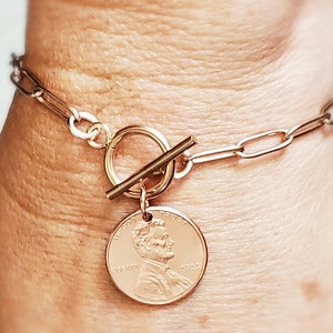 Puede incluir: Un brazalete de cadena dorada con un cierre de palanca y un dije de un centavo con el perfil de Abraham Lincoln. El dije de un centavo tiene el a&ntilde;o 2022 inscrito.