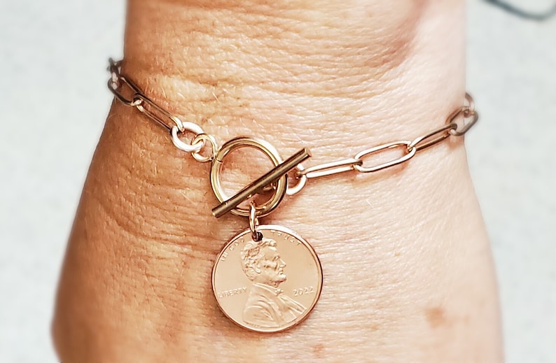 Puede incluir: Un brazalete de cadena dorada con un cierre de palanca y un dije de un centavo con el perfil de Abraham Lincoln. El dije de un centavo tiene el a&ntilde;o 2022 inscrito.