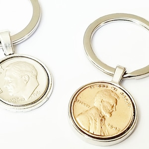 May include: Two silver keychains with a coin-shaped charm. One charm features a silver Lincoln penny with the inscription "IN GOD WE TRUST" and the year "1947". The other charm features a silver dime with the inscription "LIBERTY" and the year "1964".