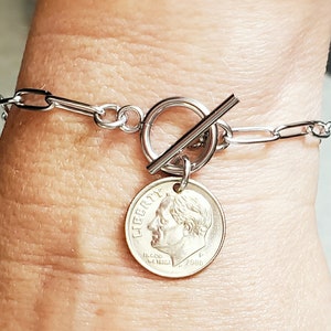 Puede incluir: Un brazalete de cadena de plata con un cierre de palanca y un dije de diez centavos de plata. La moneda de diez centavos est&aacute; fechada en 2000 y presenta el perfil del presidente Franklin D. Roosevelt.