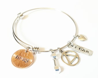 May include: Silver bangle bracelet with various charms. Charms include a copper penny with the date 10-23-2017, a dumbbell, a triangle symbol, a heart, and a rectangular charm with the text "I CAN".