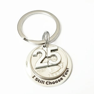 Puede incluir: Llavero plateado con un colgante redondo que presenta una moneda de veinticinco centavos y el número "25" en relieve. El texto "I Still Choose You!" está grabado alrededor de la moneda. El llavero incluye un anillo metálico.