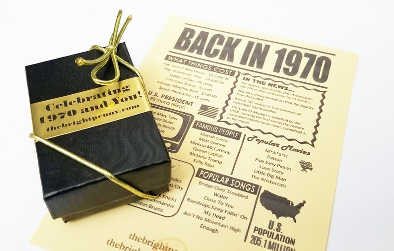 Puede incluir: Una caja de regalo negra con una cinta dorada y una etiqueta dorada que dice "Celebrating 1970 and You! thebrightpenny.com". La caja est&aacute; sobre un papel amarillo con un dise&ntilde;o de peri&oacute;dico que dice "Back in 1970" y enumera eventos, personas famosas y pel&iacute;culas populares del a&ntilde;o 1970.