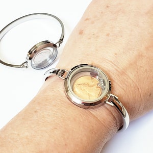 May include: Silver charm bracelet with a round glass locket containing a copper coin. The bracelet is on a person's wrist.