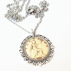 Pode incluir: Um colar com um pingente de moeda dourada e uma corrente de prata. A moeda apresenta o perfil de uma mulher e o ano de 1922. O pingente é rodeado por uma moldura decorativa de filigrana de prata.