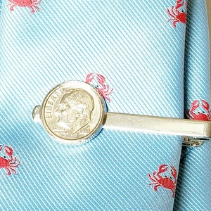 May include: A silver tie bar with a dime coin featuring the profile of President Franklin D. Roosevelt. The tie bar is on a light blue tie with a pattern of red crabs.