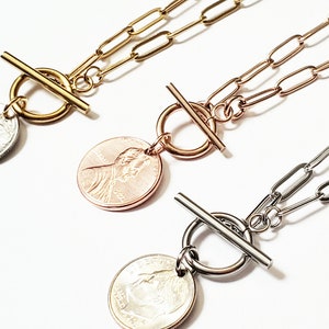May include: Three gold, rose gold, and silver necklaces with a coin pendant and a toggle clasp. The coin pendants feature a profile of a person.