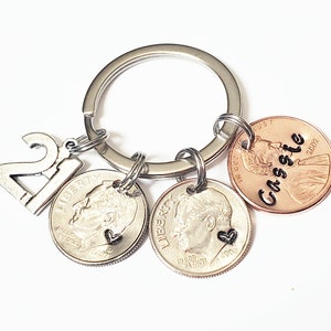May include: Silver keychain with a charm that says "21" and three coins. One coin is a US penny with the name "Cassie" engraved on it. The other two coins are US dimes with a heart charm attached to each.