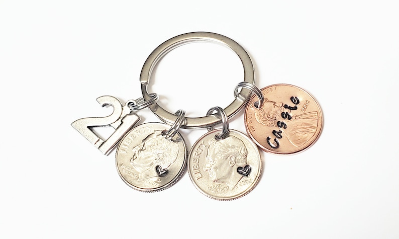 May include: Silver keychain with a charm that says "21" and three coins. One coin is a US penny with the name "Cassie" engraved on it. The other two coins are US dimes with a heart charm attached to each.