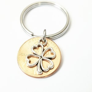 May include: A silver four-leaf clover charm attached to a penny keychain. The penny is dated 1987 and has the words "In God We Trust" and "Liberty" visible.