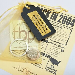 May include: A yellow organza bag with a silver key chain and three US coins. A black tag with gold lettering reads "Celebrating 2004 and You!" Thebrightpenny.com. A yellow paper with black text reads "Back in 2004" and lists popular movies and songs from 2004.