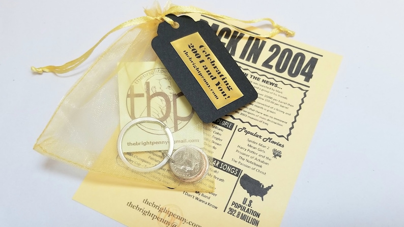 May include: A yellow organza bag with a silver key chain and three US coins. A black tag with gold lettering reads "Celebrating 2004 and You!" Thebrightpenny.com. A yellow paper with black text reads "Back in 2004" and lists popular movies and songs from 2004.