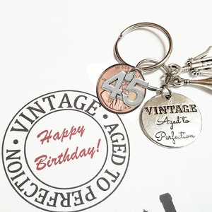 May include: A silver keyring with a silver charm that says "Vintage Aged to Perfection" and a penny with the number "45" on it. The keyring also has a silver bottle opener and a silver bottle charm.