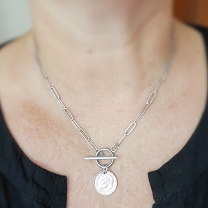 Puede incluir: Un collar de cadena de plata con un colgante de moneda de plata y un cierre de palanca. La moneda tiene un dise&ntilde;o en relieve.