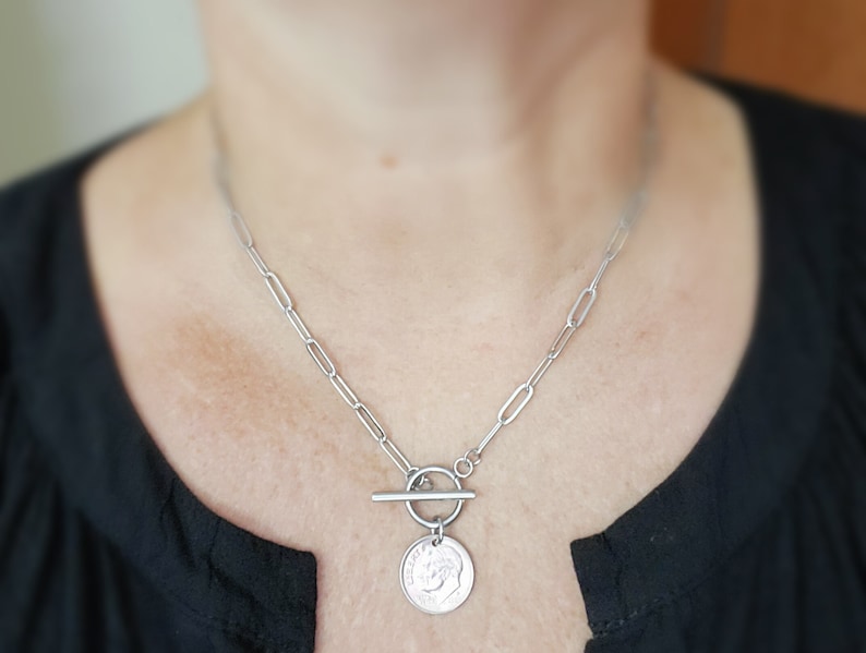 Puede incluir: Un collar de cadena de plata con un colgante de moneda de plata y un cierre de palanca. La moneda tiene un dise&ntilde;o en relieve.