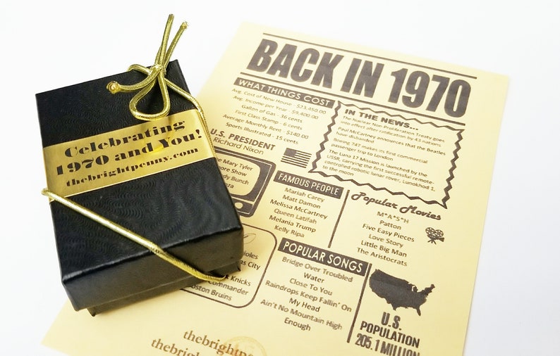 May include: A black gift box with a gold ribbon and a gold label that says "Celebrating 1970 and You! thebrightpenny.com". The box is sitting on a yellow paper with a newspaper-style design that says "Back in 1970" and lists events, famous people, and popular movies from the year 1970.