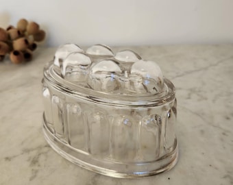 May include: Clear glass jelly mold with a domed top and an oval shape. The mold features a series of vertical indentations around the sides and a series of rounded domes on the top. The glass is transparent, allowing light to pass through.