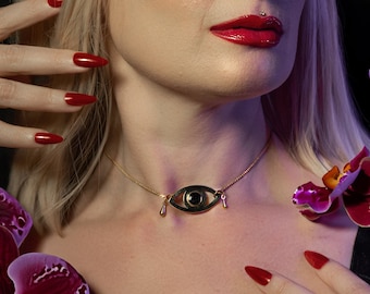 May include: A woman with blonde hair and red nail polish wears a gold necklace with a black eye pendant. She is surrounded by purple orchids.