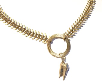 May include: Gold-toned necklace featuring a fishbone-like chain. A circular pendant with a tooth charm hangs from the center. The necklace has a lobster clasp closure.