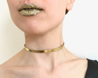 May include: A gold choker necklace with a small circle pendant. The necklace is made of a thin, flat band of metal.