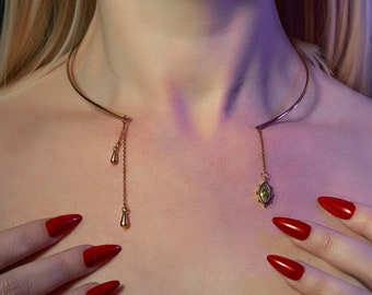 May include: A gold choker necklace with a delicate chain and two teardrop pendants. The necklace is worn by a person with red nail polish and a lip piercing.