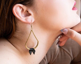 May include: Gold-toned teardrop earrings with a black leather bow detail. The earrings are suspended from a delicate chain and feature a curved, open design. The image shows the earrings worn by a person.