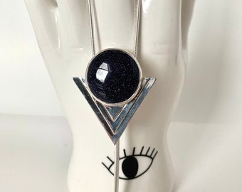 May include: A silver necklace featuring a dark blue, round stone pendant set in a silver frame. The pendant is suspended from a delicate silver chain and is displayed on a white ceramic hand.