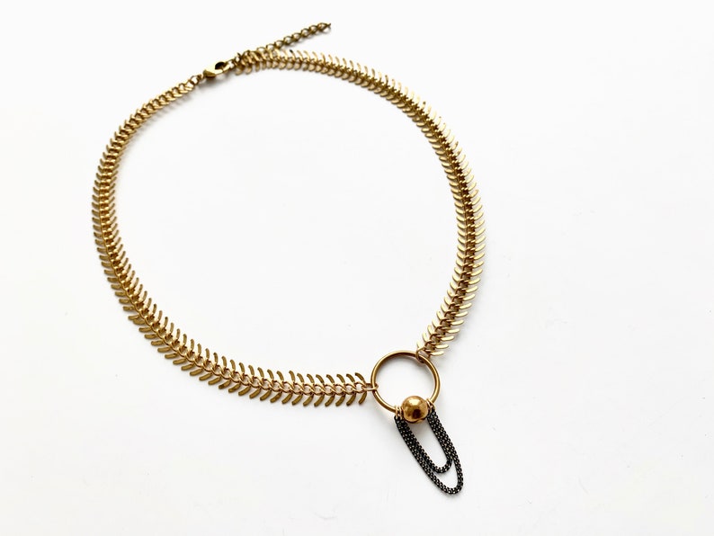 May include: A gold necklace with a unique, spiky chain design. The necklace features a dangling charm with a gold ring and a small gold ball attached to two black chains.