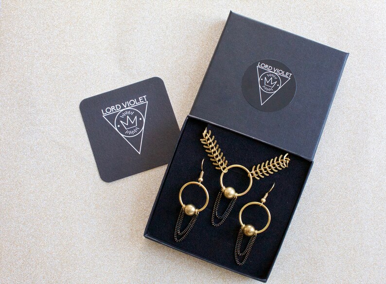 May include: A black box with a gold necklace and earrings. The box has the logo "LORD VIOLET" with a crown on it.