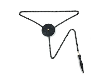 May include: A black bolo tie with a circular pendant featuring a gold center. The tie has a black cord with a pointed tip.