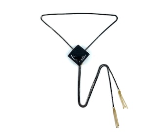 May include: A black bolo tie with a square, glossy pendant. The tie features a black chain and gold-colored tassel ends. The jewelry is set against a white background.