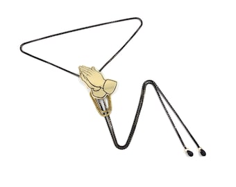 May include: A gold-colored praying hands bolo tie with a black chain. The chain is made of two strands and has a black tassel at the end.